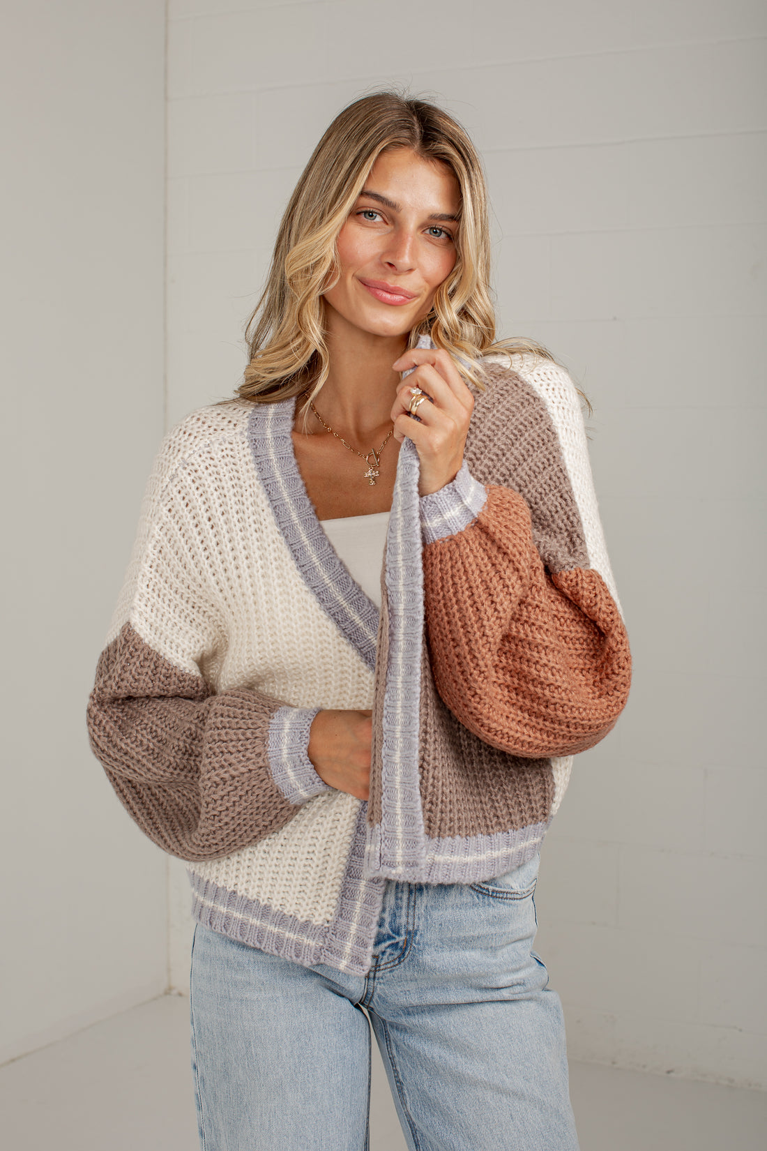 A woman wearing a chunky knit color-block cardigan with cream, taupe, and terracotta sections, paired with light-wash blue jeans, standing against a light gray wall.
