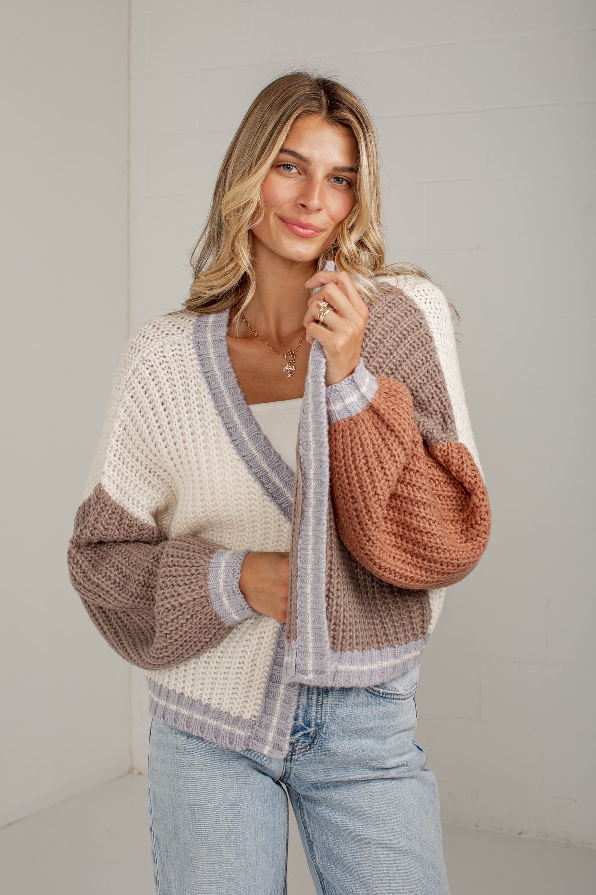 A woman wearing a chunky knit color-block cardigan with cream, taupe, and terracotta sections, paired with light-wash blue jeans, standing against a light gray wall.