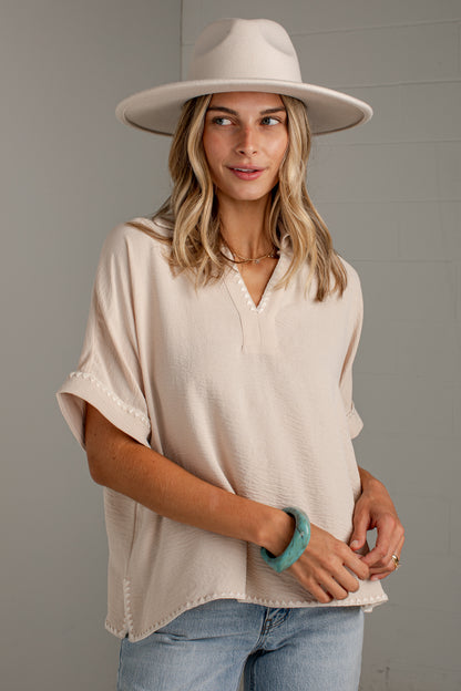 A woman wearing a light beige short-sleeve blouse with a split V-neck and scalloped edges, paired with light blue jeans and a wide-brimmed cream hat, standing against a gray wall.