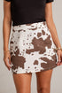 A woman modeling a black scoop-neck top and a white mini skirt with a brown cow-print pattern, styled with a cream wide-brim hat and cream ankle boots against a white studio wall.