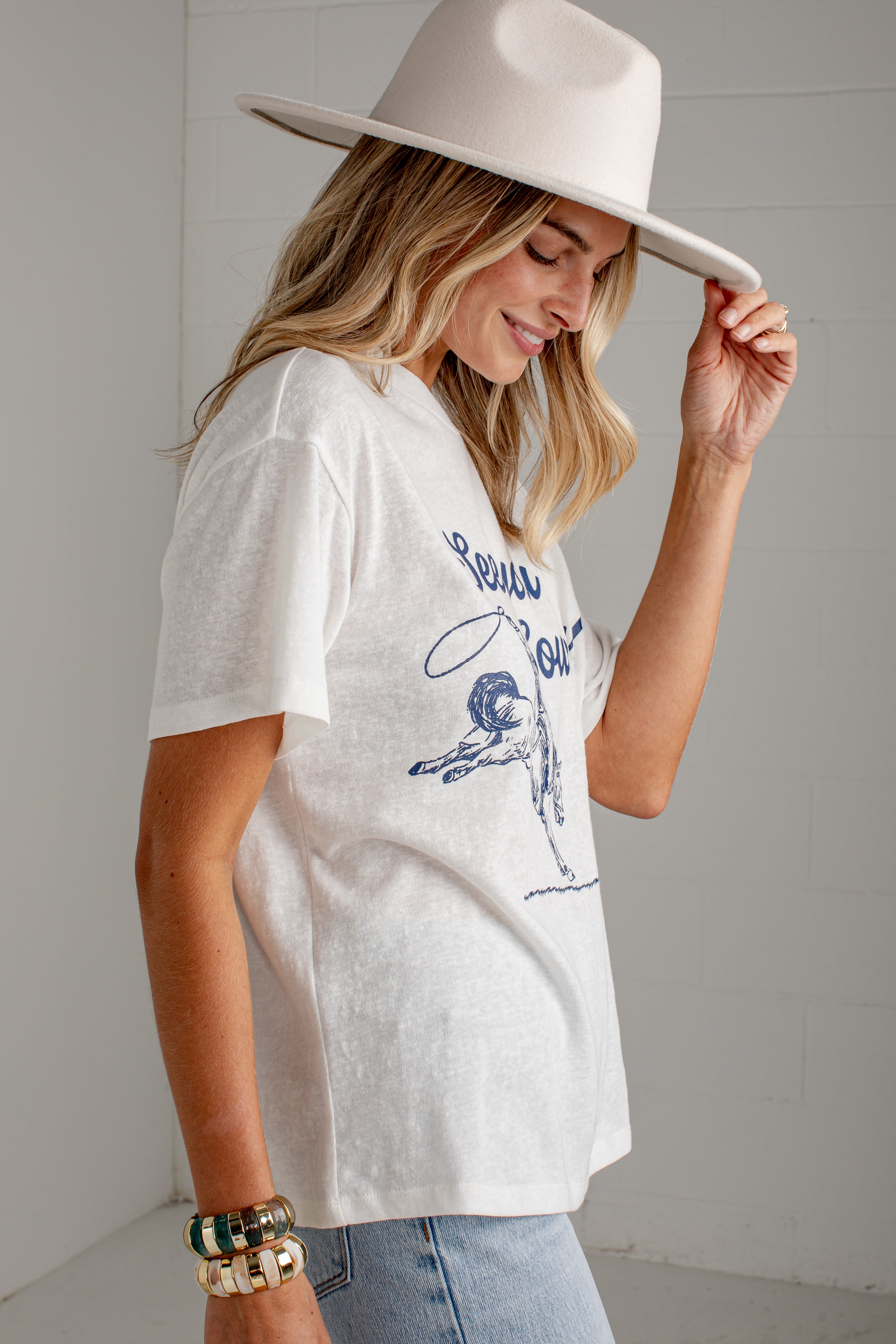 A woman wearing a white graphic t-shirt with a blue vintage-style print, paired with light-wash jeans and a wide-brimmed cream hat, posing in a bright, modern room.
