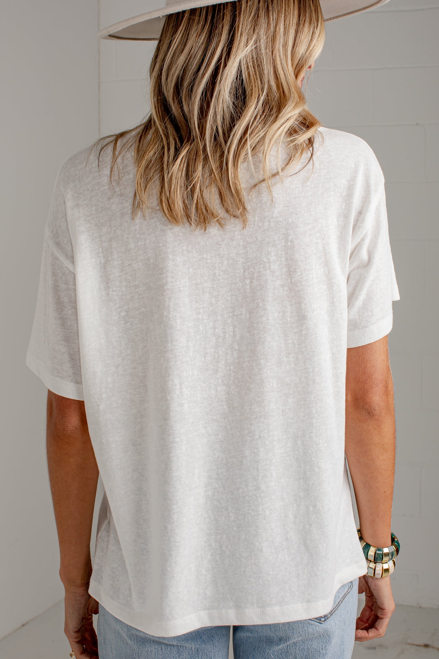 A woman viewed from behind wearing an oversized off-white short-sleeve t-shirt, light blue jeans, and a wide-brimmed light-colored hat, set against a neutral light gray background.