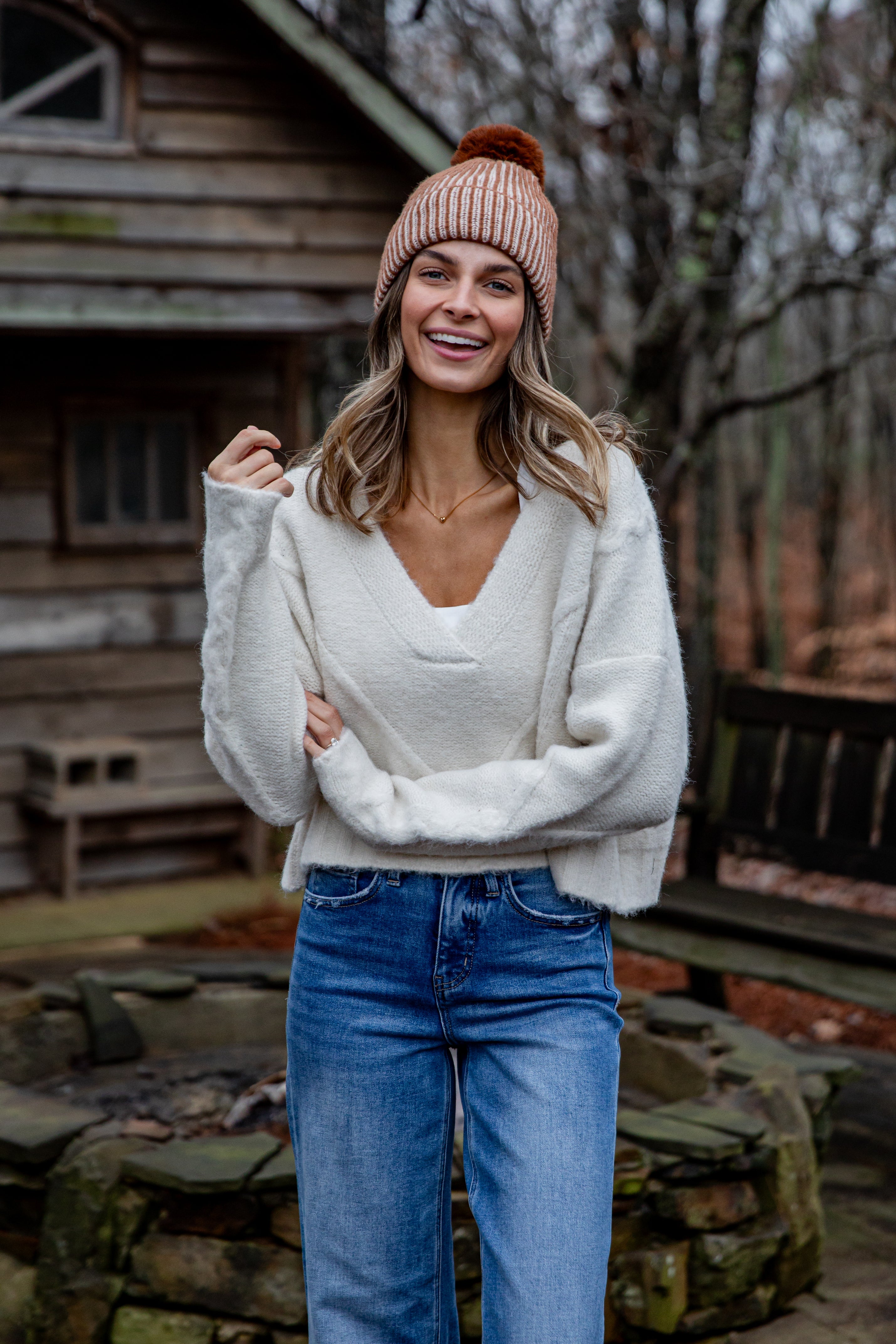 Woman wearing a white sweater and blue jeans standing in front of a wooden cabin.