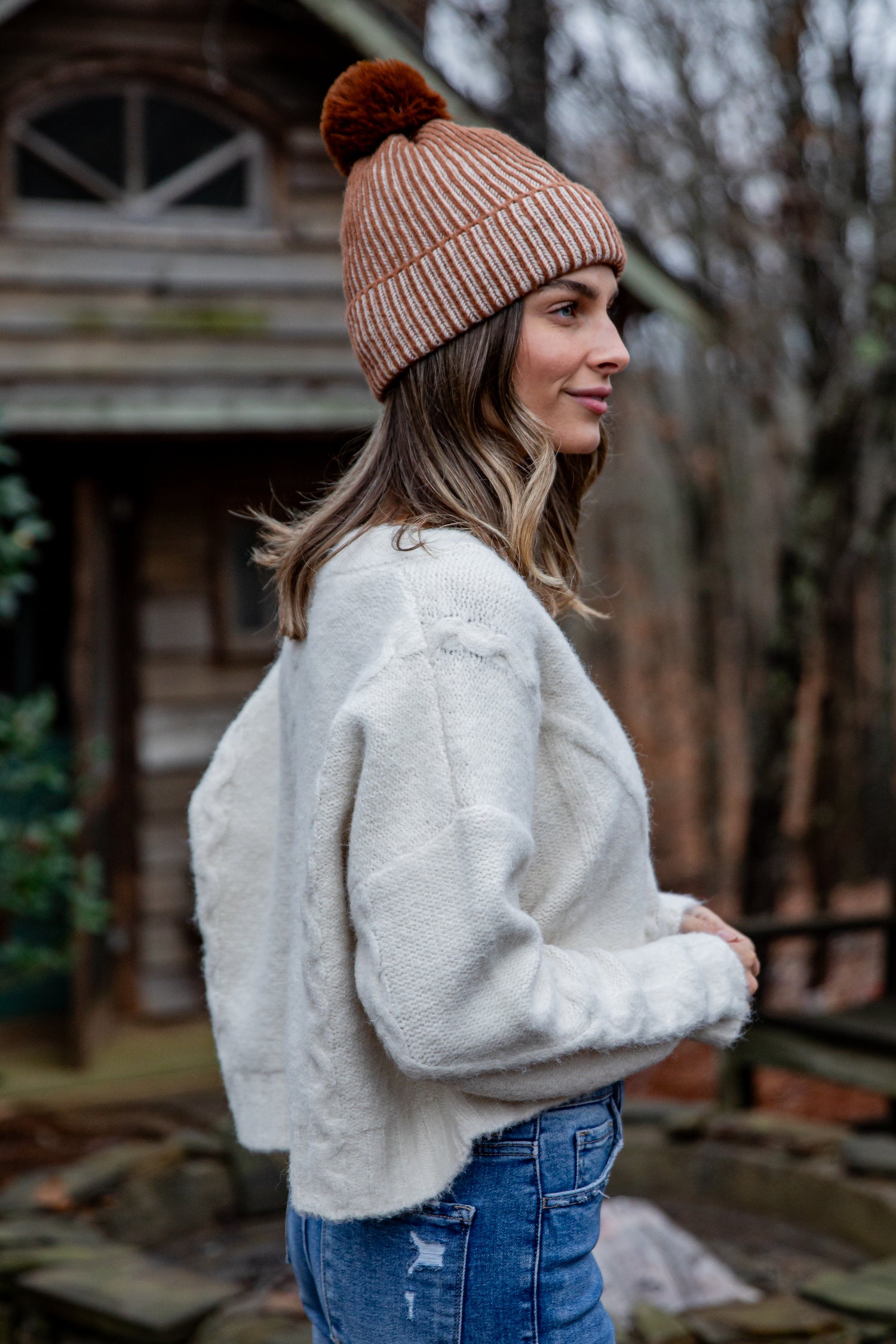 Woman wearing a beige knit beanie with a pom-pom and a white sweater outdoors.