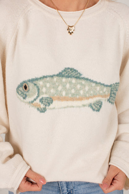A close-up of a cream-colored knit sweater featuring a knitted fish graphic in shades of teal, green, and beige, worn by a model.