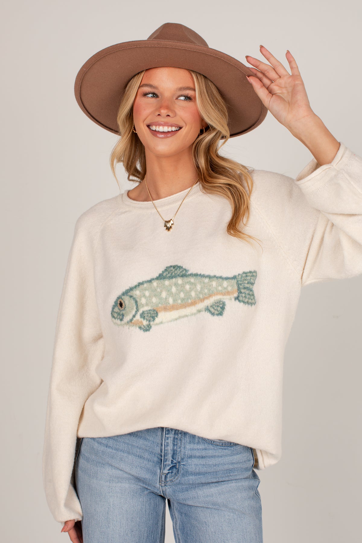 A woman wearing a cream-colored knit sweater with a fish graphic, blue jeans, and a brown wide-brim hat, posing against a light gray studio background.