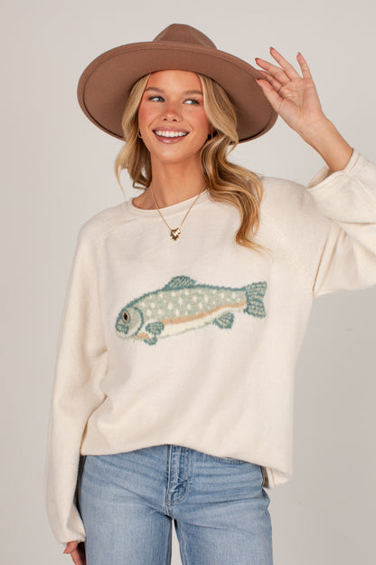 A woman wearing a cream-colored knit sweater with a fish graphic, blue jeans, and a brown wide-brim hat, posing against a light gray studio background.