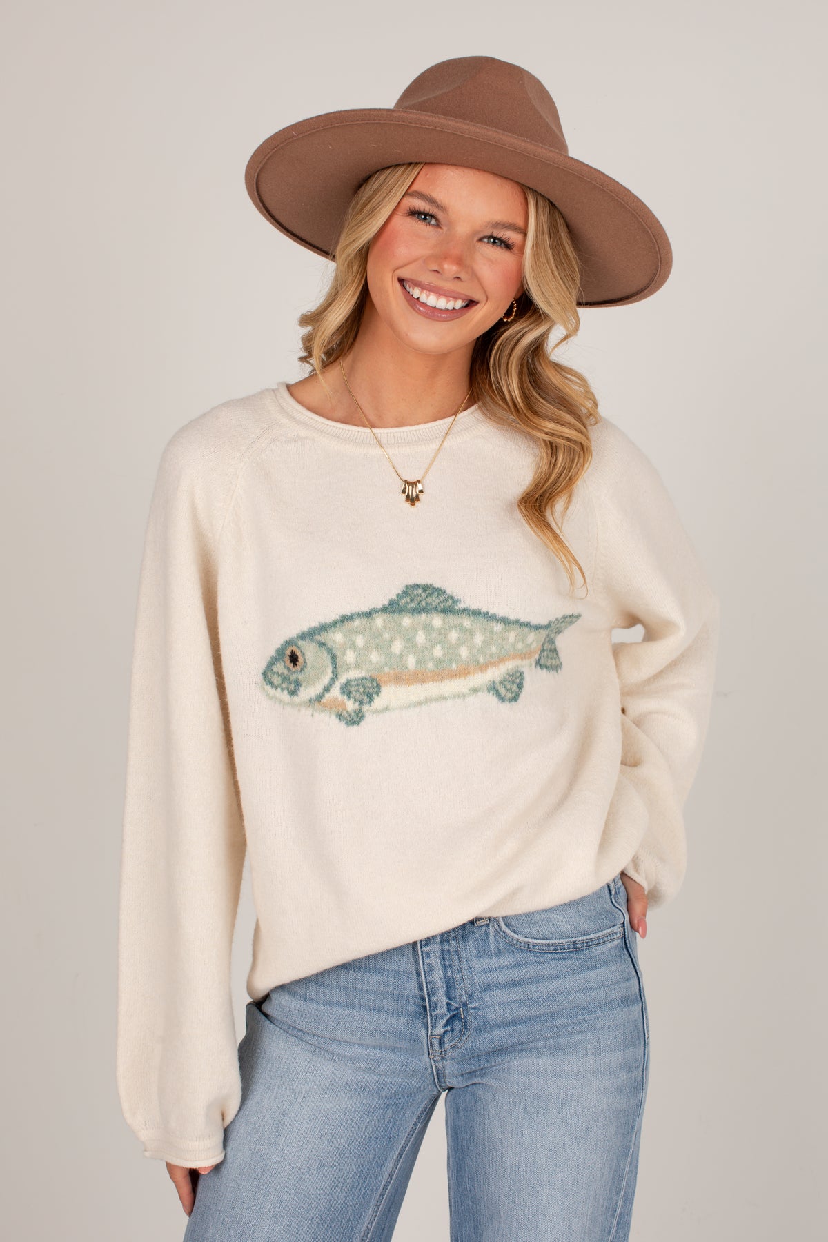 A smiling blonde woman wearing a cream-colored knit sweater with a detailed fish graphic, blue jeans, and a brown wide-brim hat, photographed against a neutral light gray studio background.