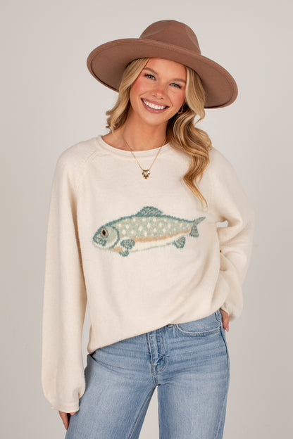 A smiling blonde woman wearing a cream-colored knit sweater with a detailed fish graphic, blue jeans, and a brown wide-brim hat, photographed against a neutral light gray studio background.