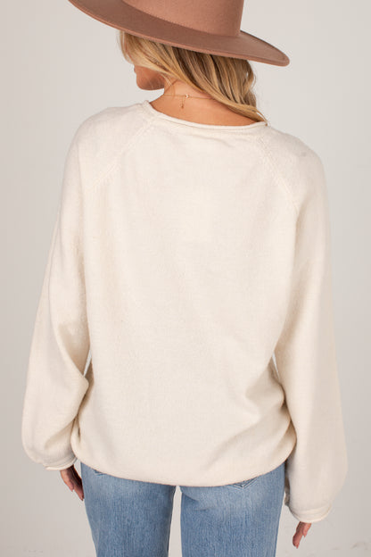 A woman shown from the back wearing a cream-colored long-sleeve knit sweater, blue jeans, and a brown wide-brim hat against a light gray studio background.