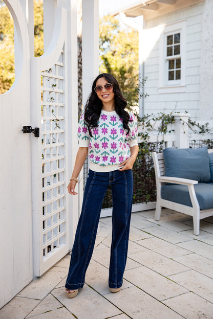 Woman standing outdoors on a patio wearing a floral top and blue jeans.