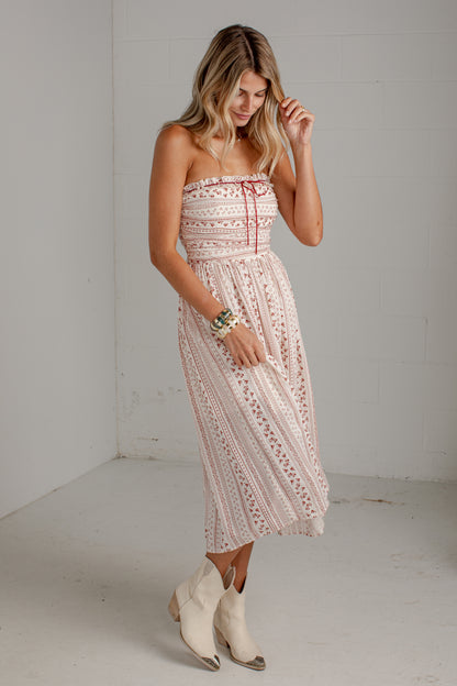 A woman modeling a strapless midi dress with a cream and pink bohemian lace pattern, paired with cream western boots, in a minimalist white room.