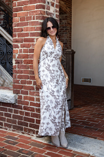 A woman wearing a sleeveless white halter maxi dress with a gray floral pattern and gray boots, standing outdoors next to a red brick column.