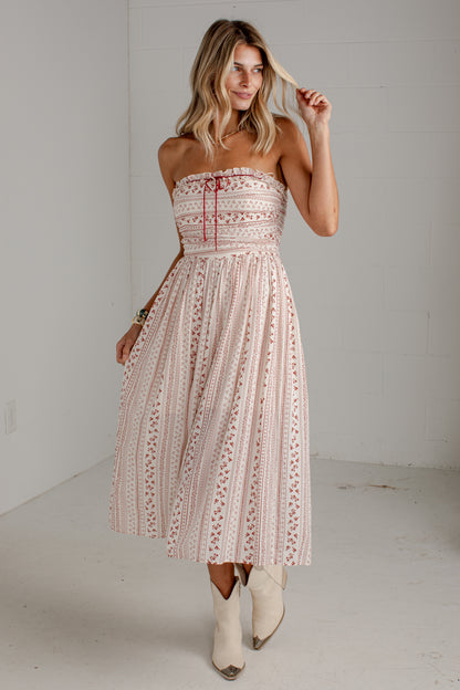 A woman modeling a strapless midi dress with a light pink and white bohemian-style pattern, featuring a red drawstring at the bust. She is wearing off-white cowboy boots in a bright, neutral indoor studio setting.