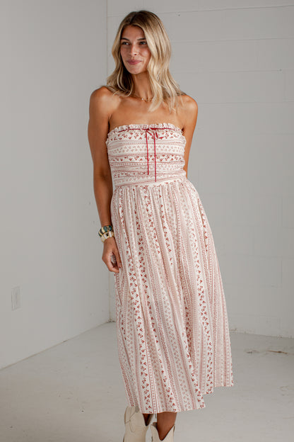 A woman modeling a strapless midi dress with a light pink and white bohemian-style pattern and red tie details, standing against a white studio wall.