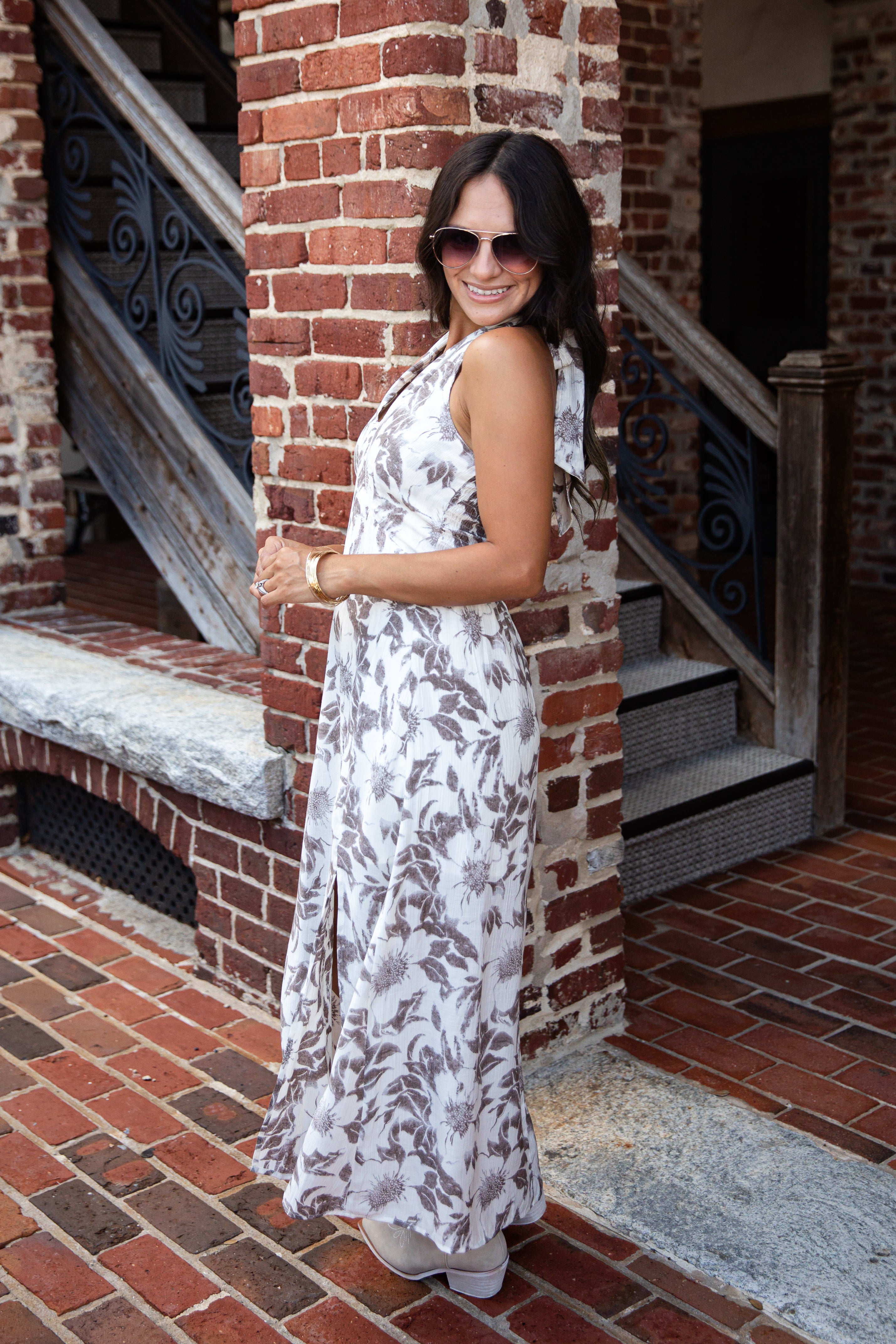 A woman wearing a sleeveless white maxi dress with a brown floral pattern, standing on a brick patio in front of a brick building with wooden railings.