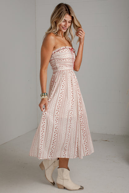 A woman modeling a strapless midi dress with a light pink and white bohemian-style geometric pattern, paired with cream-colored cowboy boots, in a bright studio setting.