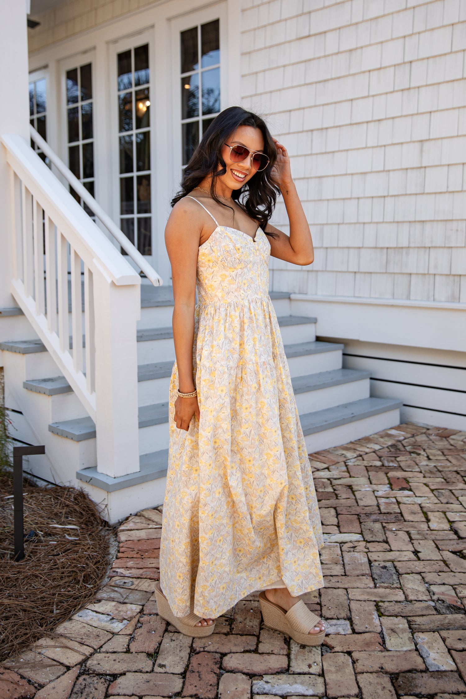 Woman in a yellow floral dress standing on a stone path in front of a white house.
