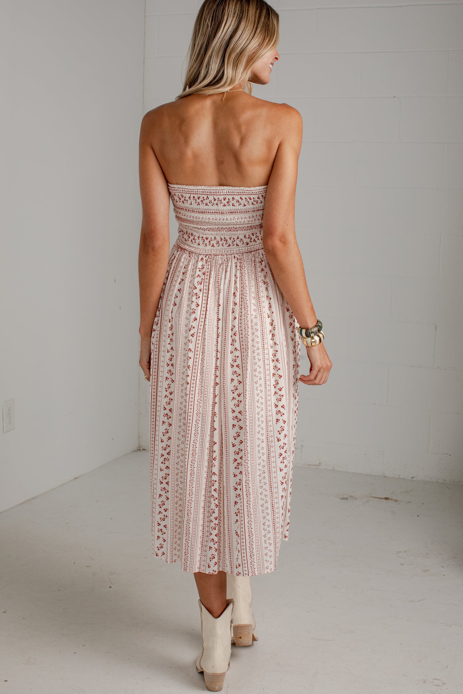 A woman shown from behind wearing a strapless midi dress with a pleated, embroidered lace-like pattern in a dusty pink color, paired with beige cowboy boots in a white studio setting.