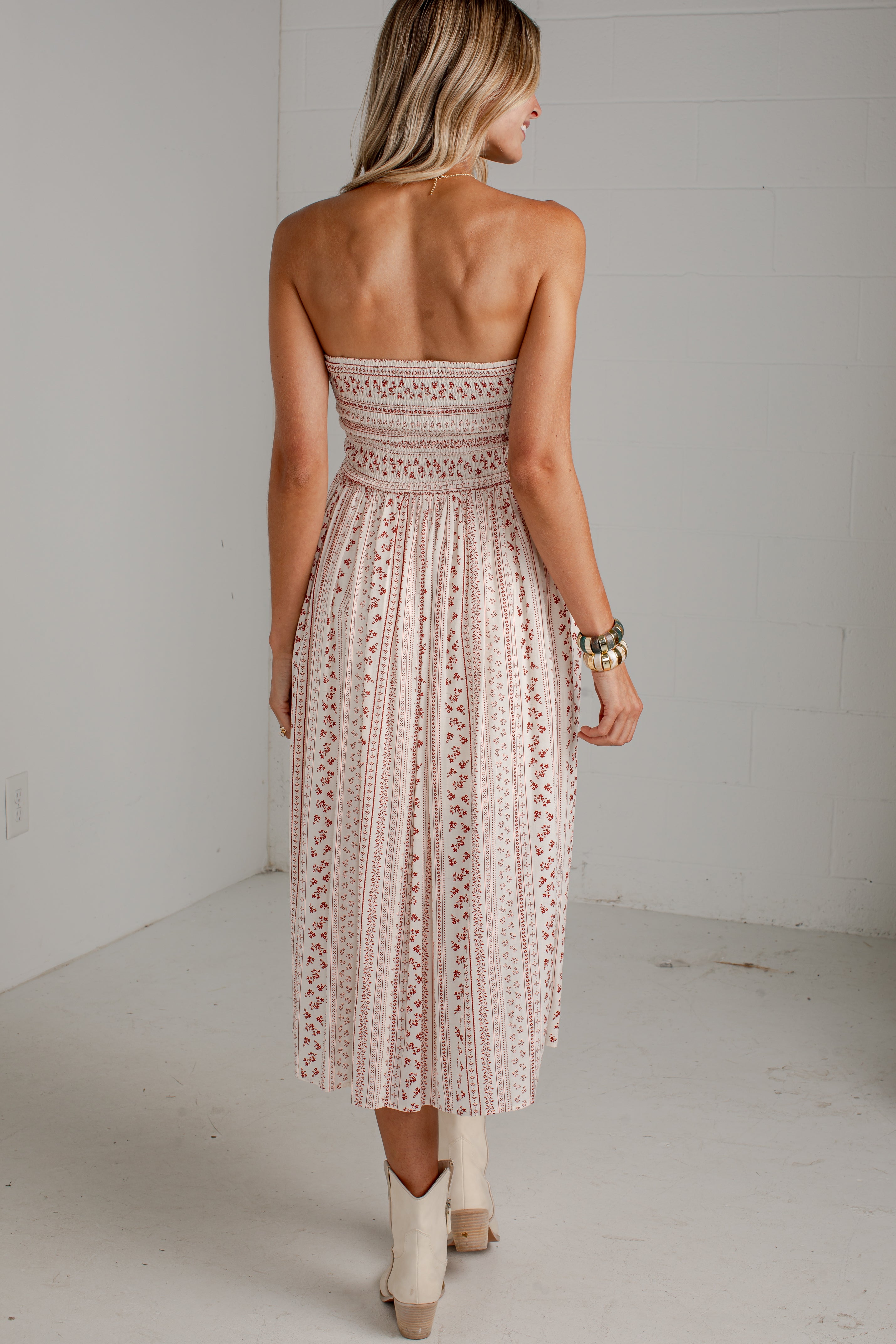 A woman shown from behind wearing a strapless midi dress with a pleated, embroidered lace-like pattern in a dusty pink color, paired with beige cowboy boots in a white studio setting.
