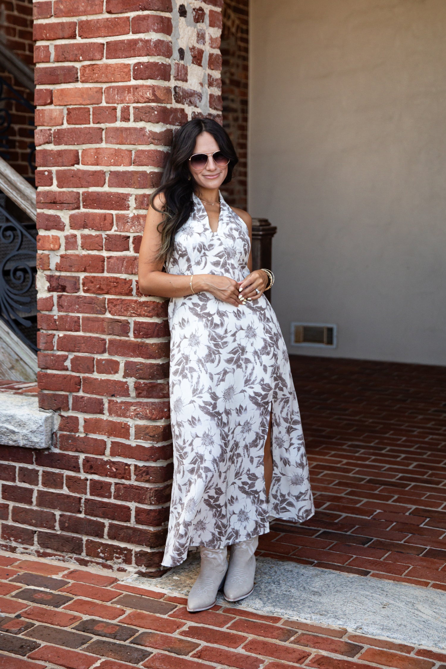 A woman wearing a sleeveless white halter maxi dress with a gray botanical leaf pattern and gray boots, posing outdoors against a red brick column.