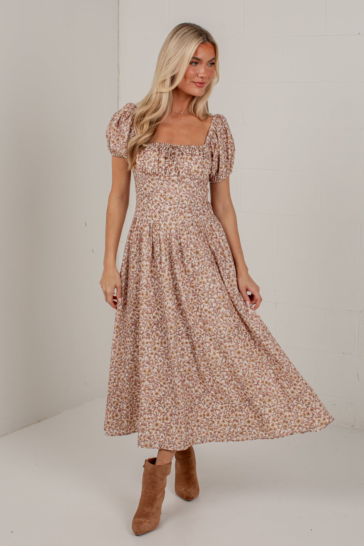 A woman modeling a midi-length puff-sleeve dress with a small floral print in beige, brown, and muted pink tones, paired with tan ankle boots, set against a plain off-white studio background.