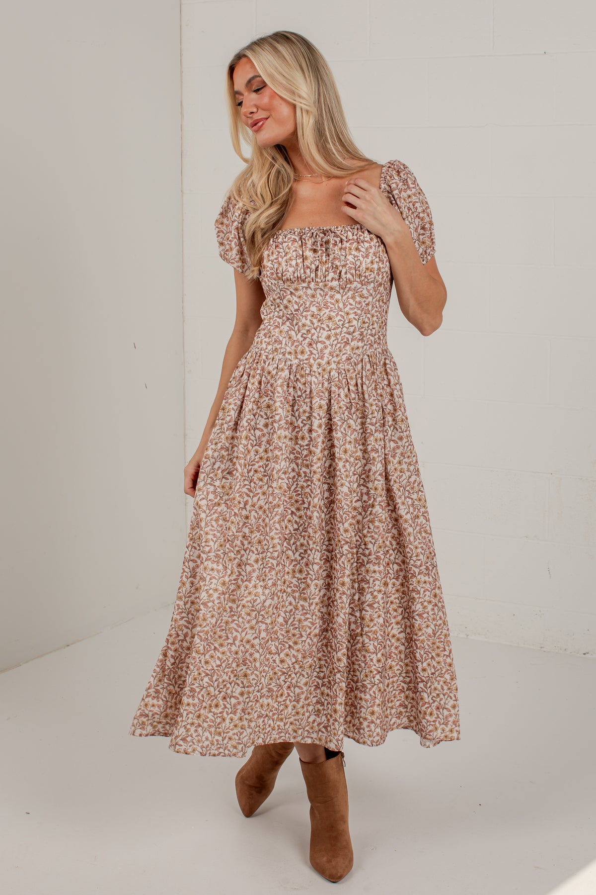 A woman modeling a midi-length puff-sleeve dress with a small beige and brown floral print, paired with brown ankle boots, set against a plain white studio background.