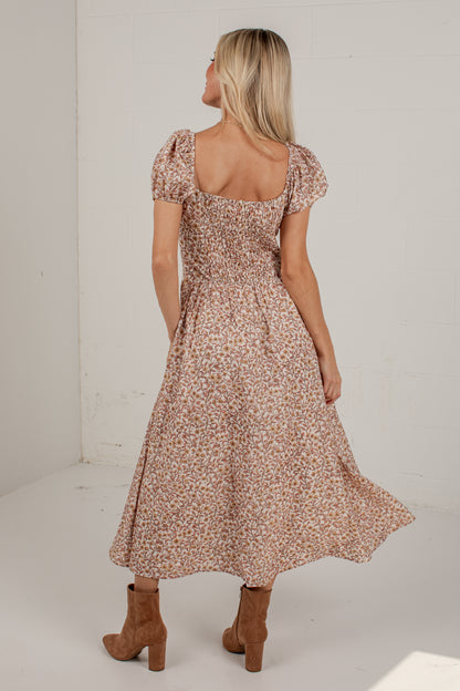 A woman wearing a brown and beige floral print midi dress with puff sleeves and a smocked bodice, shown from the back in a bright studio setting.