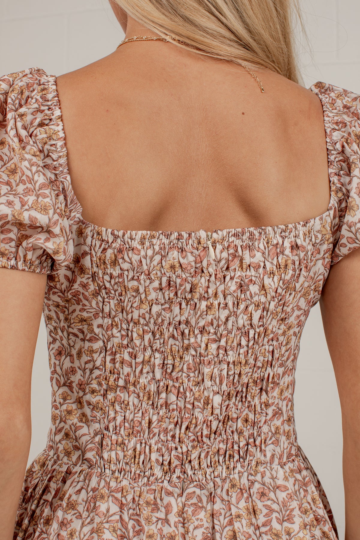 Close-up back view of a woman wearing a smocked floral dress with puff sleeves and a square neckline. The dress has a warm beige base with brown and muted orange floral print.
