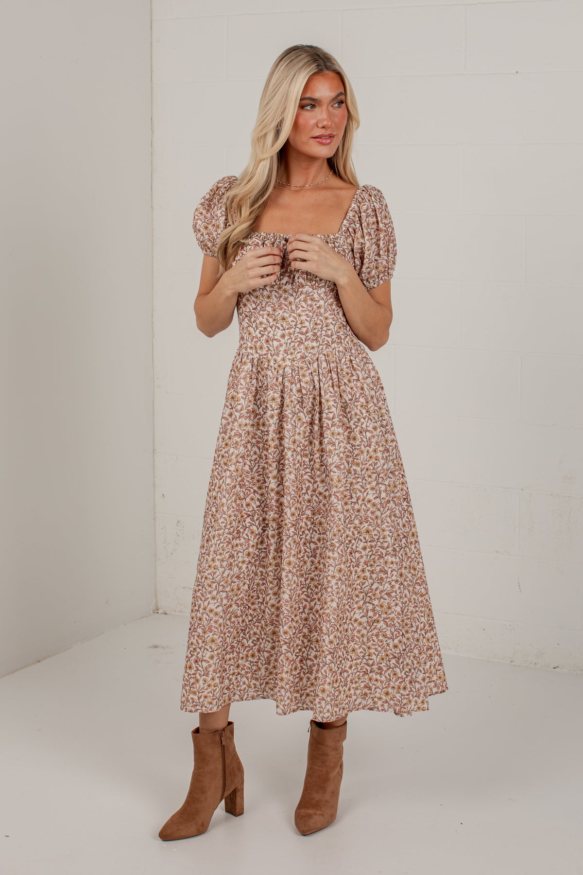 A woman modeling a midi-length puff-sleeve dress with a small-scale floral print in beige, brown, and muted pink tones, paired with brown suede ankle boots, set against a plain white studio background.