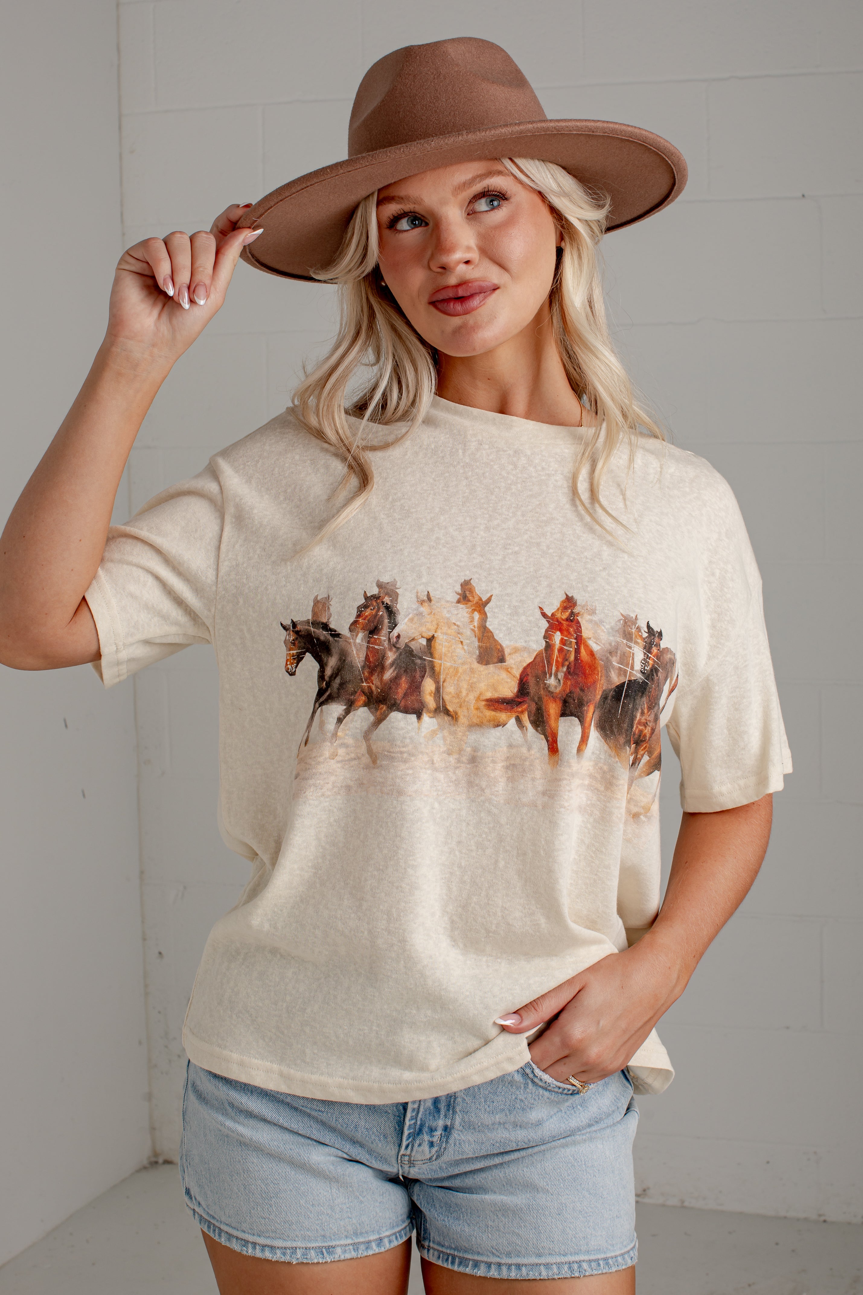 A woman wearing a cream-colored short-sleeve t-shirt with a graphic print of a herd of horses, paired with denim shorts and a brown wide-brim hat, posing against a light gray wall.