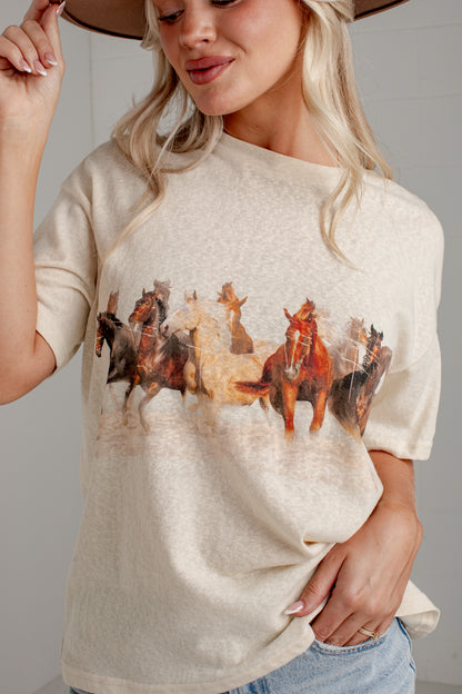 A woman wearing a cream-colored short-sleeve t-shirt with a graphic print of running horses, paired with light blue jeans and a brown hat.