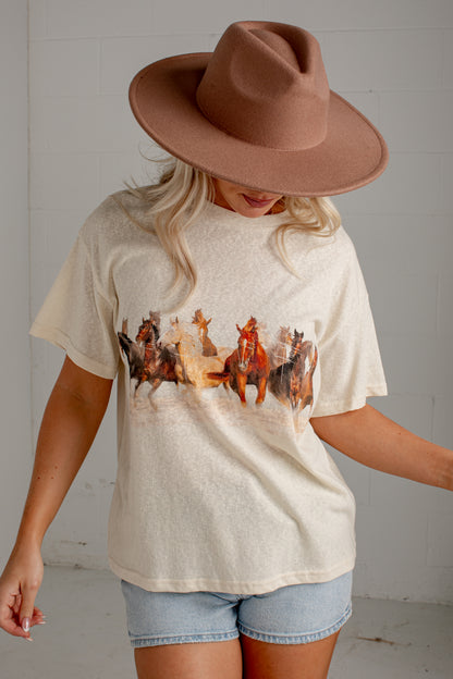 A woman wearing a cream-colored short-sleeve t-shirt with a western-themed horse and rider graphic, paired with denim shorts and a brown wide-brim hat, standing against a white brick wall.