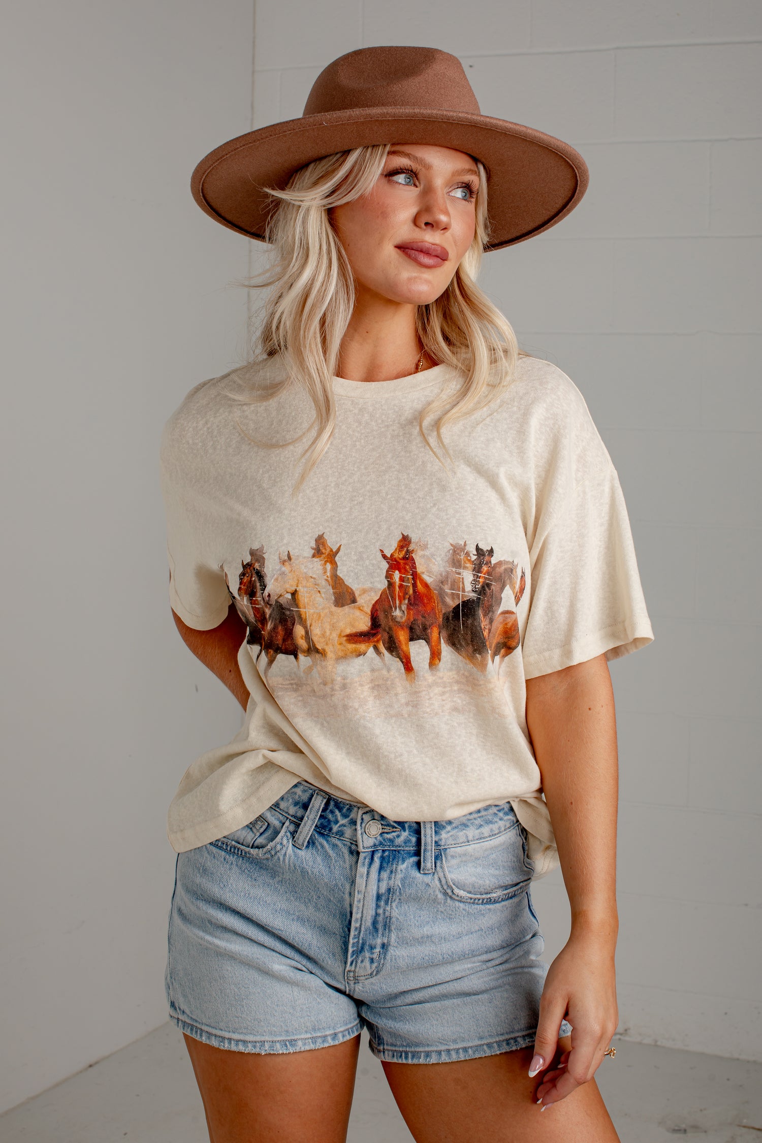 A woman wearing a cream-colored graphic t-shirt with a western horse print, paired with denim shorts and a brown wide-brim hat, standing against a light gray wall.