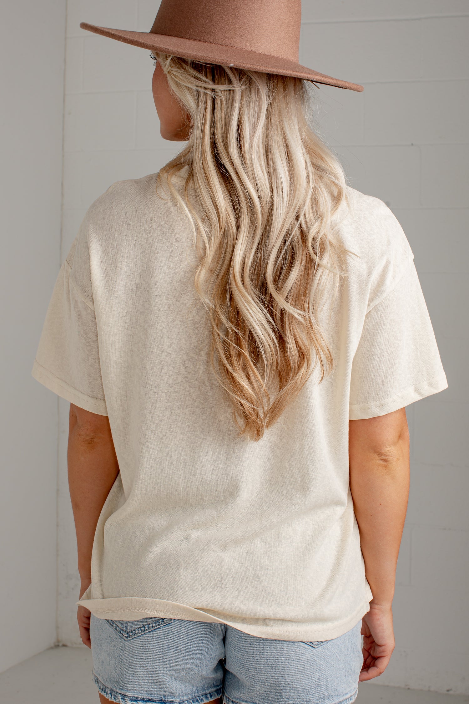 A woman viewed from behind wearing a cream-colored short-sleeve t-shirt, light-wash denim shorts, and a brown wide-brim hat against a white brick wall.