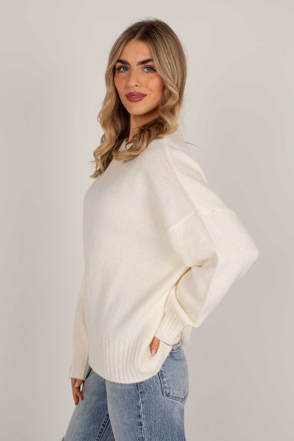 A woman wearing a cream-colored knit sweater and light blue jeans, posing against a neutral light gray studio background.