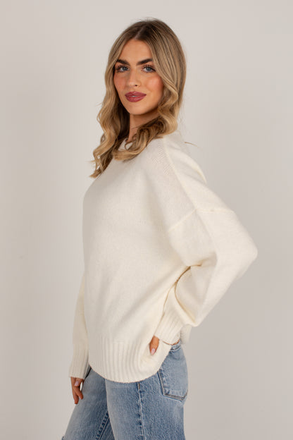 A woman wearing a cream-colored knit sweater and light blue jeans, posing against a neutral light gray studio background.