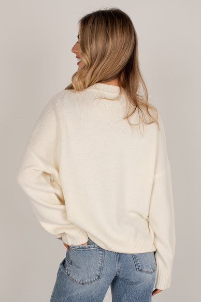 A woman with blonde hair is shown from the back wearing a cream-colored knit sweater and blue jeans against a light gray studio background.