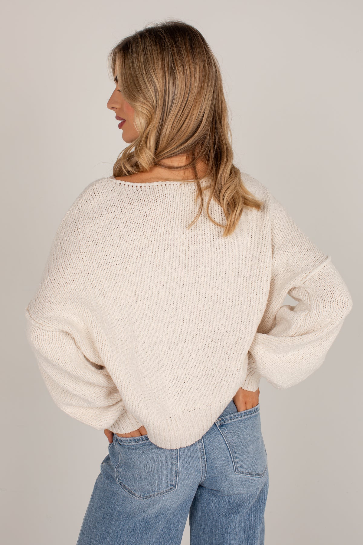 A woman shown from the back wearing a cream-colored, textured knit sweater and light blue jeans against a plain light gray studio background.