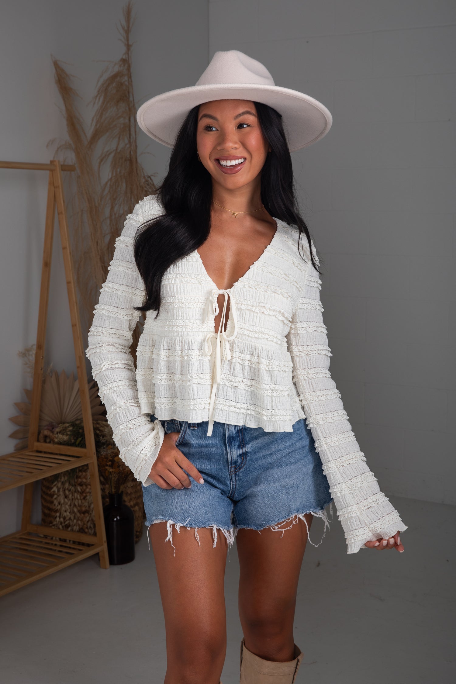 Woman wearing a white lace top and denim shorts with a white hat indoors.