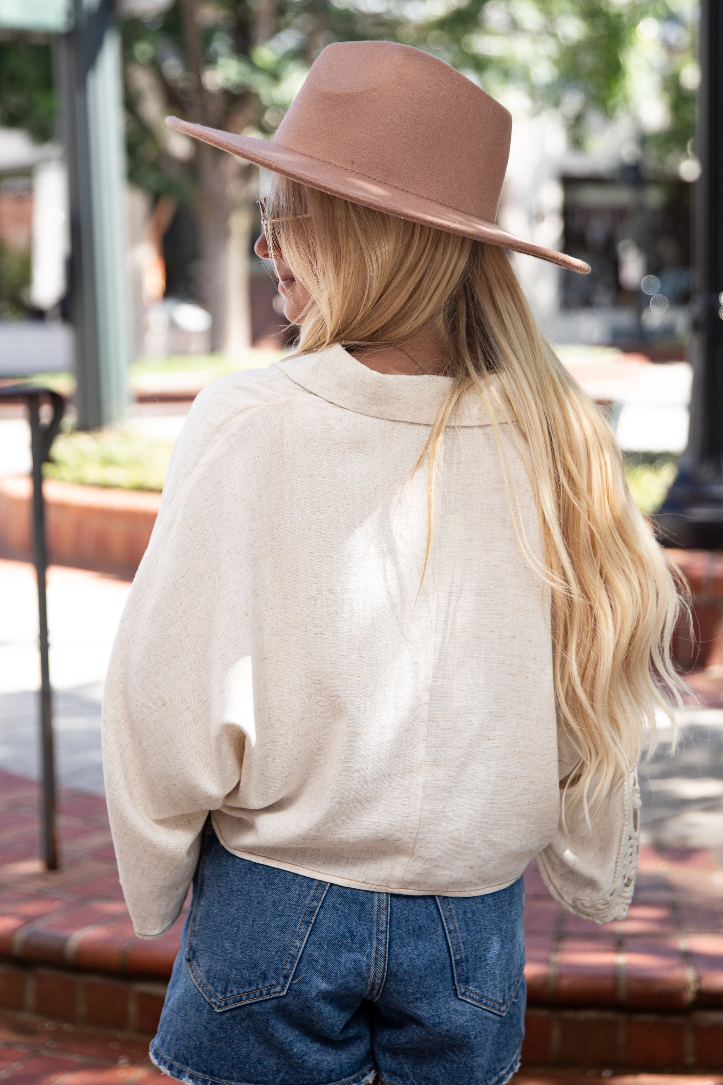 A woman photographed from behind outdoors, wearing a light beige oversized long-sleeve top, blue denim shorts, and a dusty pink wide-brim fedora hat.