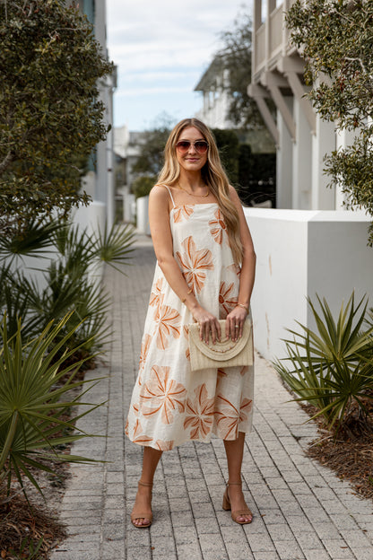 Woman in a floral dress standing on a sidewalk with plants and buildings in the background