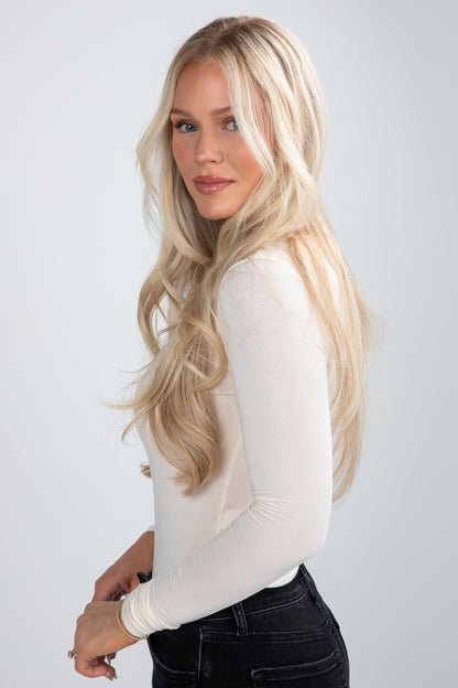 Woman with long blonde hair wearing a white long-sleeve top against a plain background