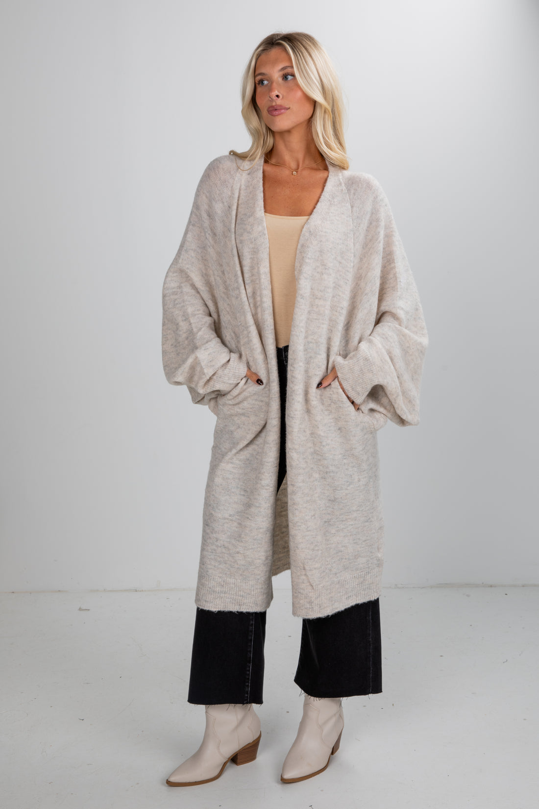 Woman wearing a long beige cardigan with black pants and beige boots on a white background