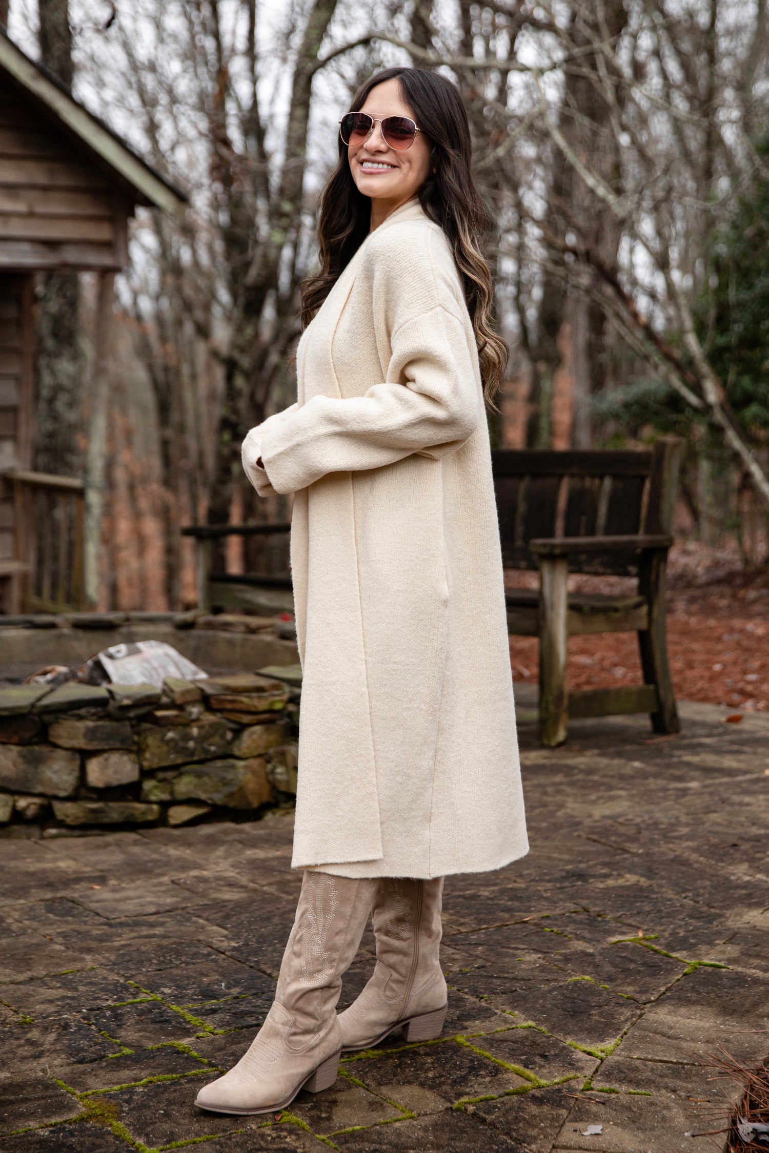 Woman wearing a long beige coat and knee-high boots standing outdoors in a wooded area.