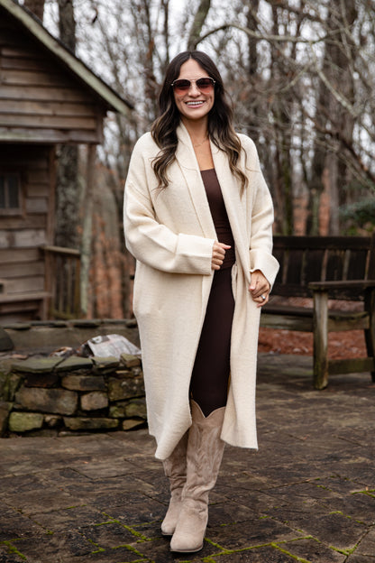 Woman wearing a long cream coat in an outdoor setting with bare trees and a wooden cabin.