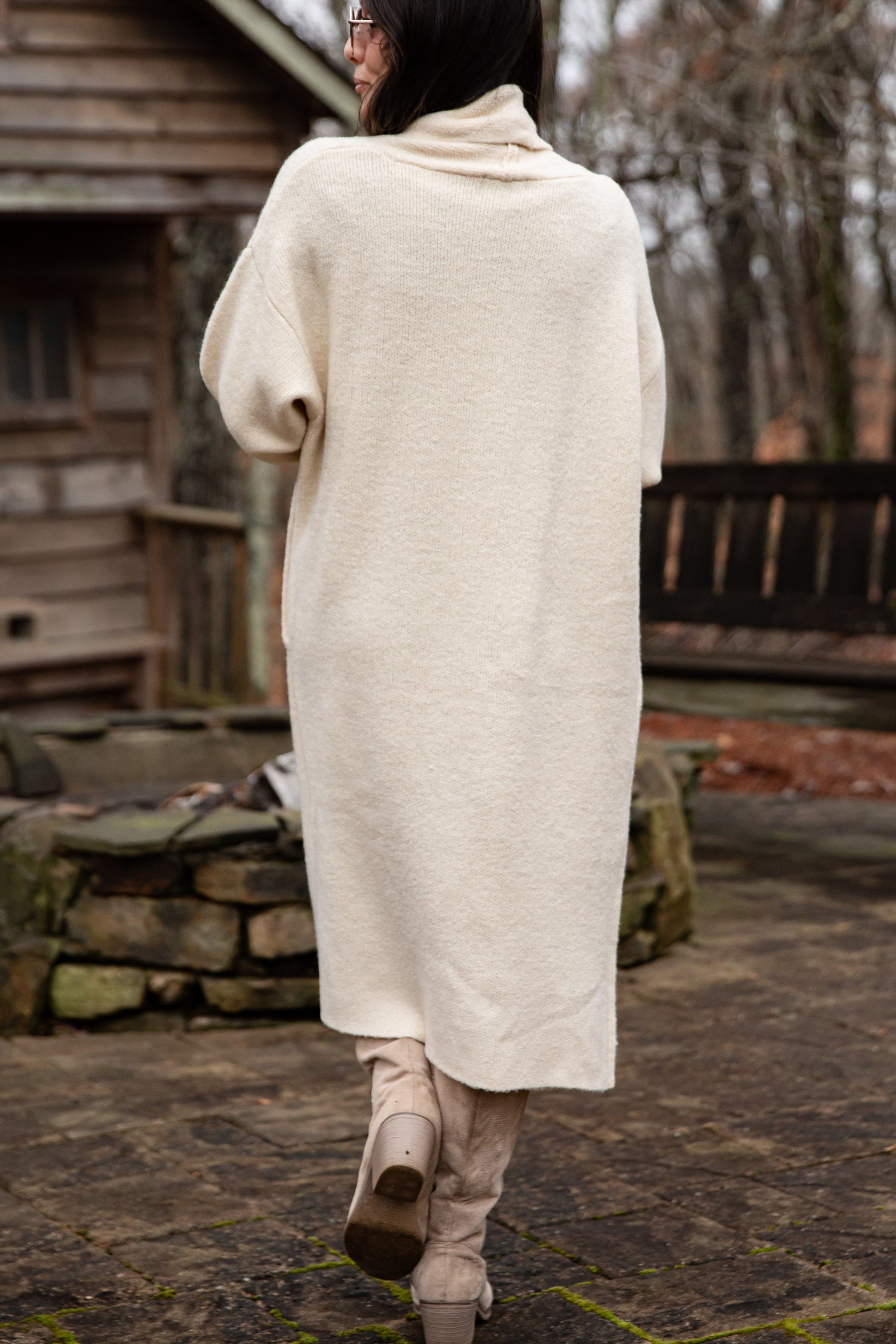 Person wearing a long cream coat walking outdoors near a wooden cabin.