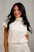 A smiling woman with long dark hair wearing a cream-colored knit turtleneck sweater vest and a matching pleated skirt, set against a light gray studio background.