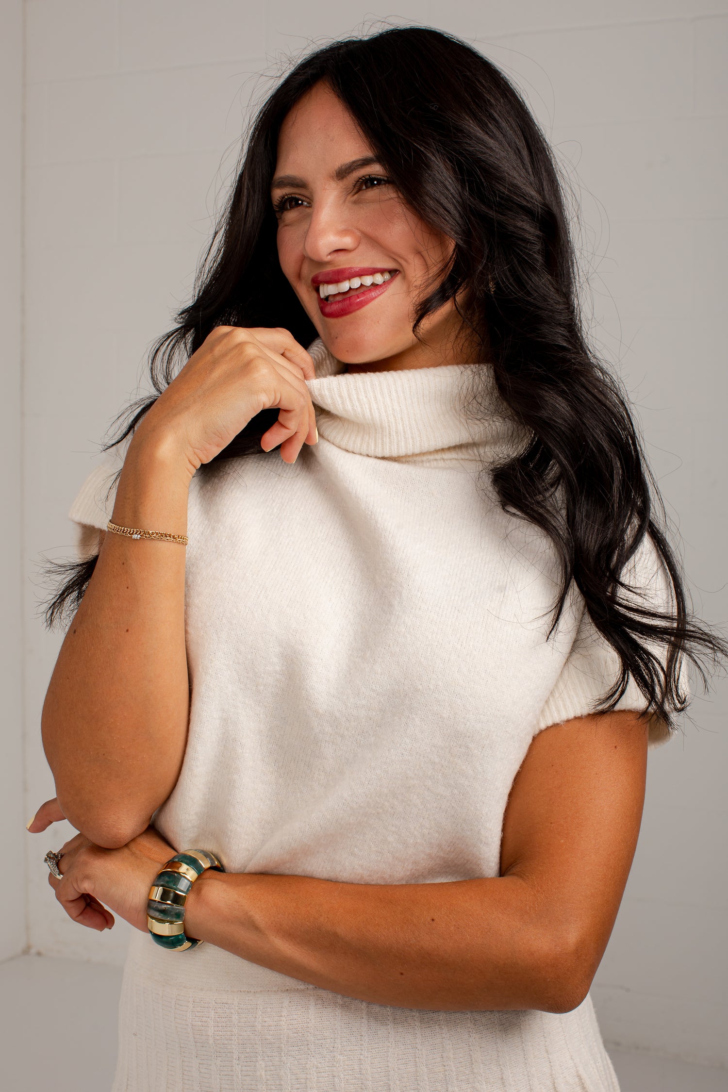 A smiling woman with long dark hair wearing a cream-colored short-sleeve knit turtleneck sweater, posing against a light off-white background.