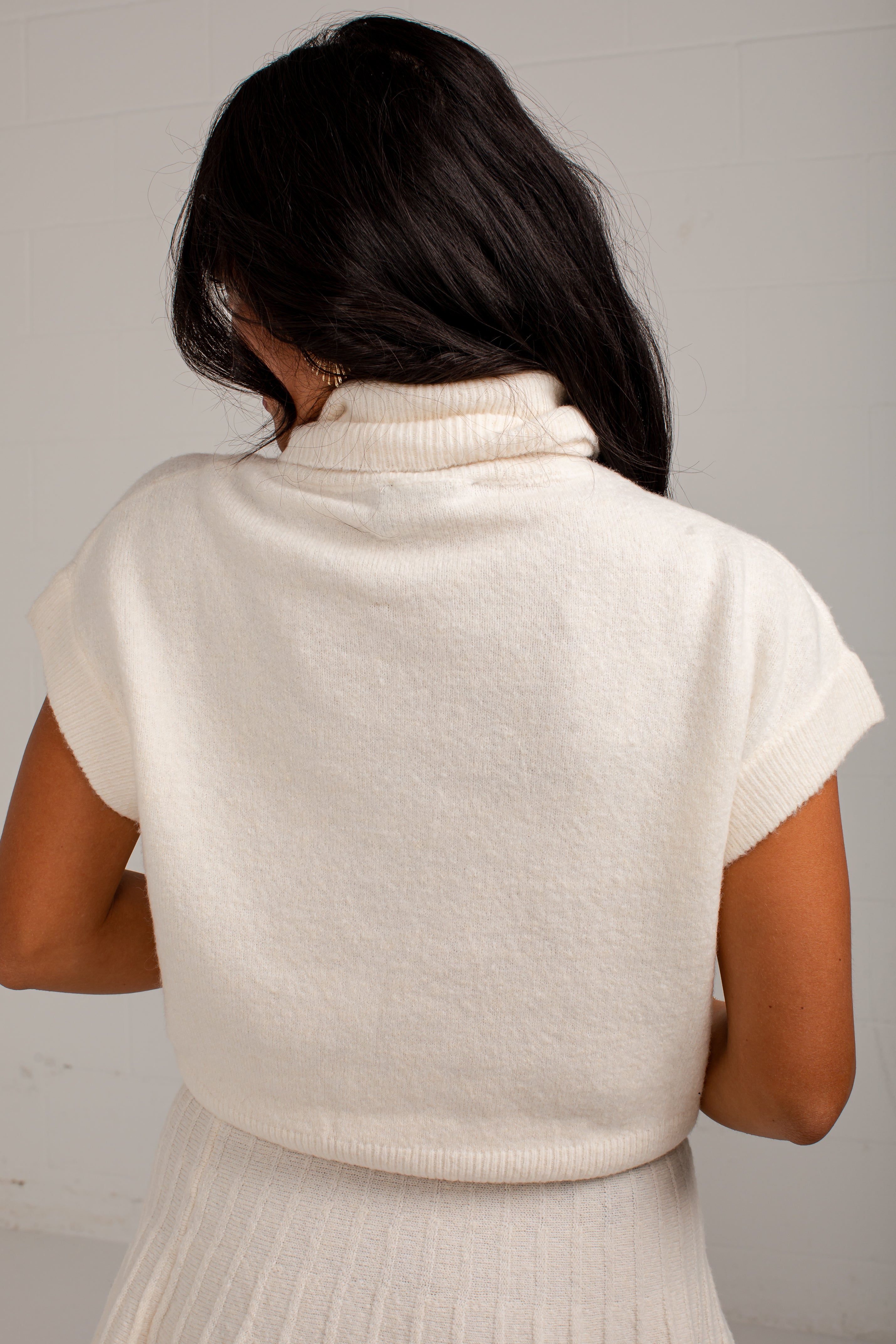 Back view of a woman wearing a cream-colored short-sleeve knit turtleneck sweater and a matching cream pleated skirt against a light gray wall.